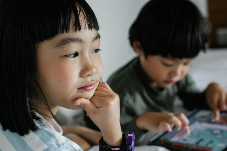 Crop concentrated ethnic girl touching chin while sitting near sibling playing game in modern device