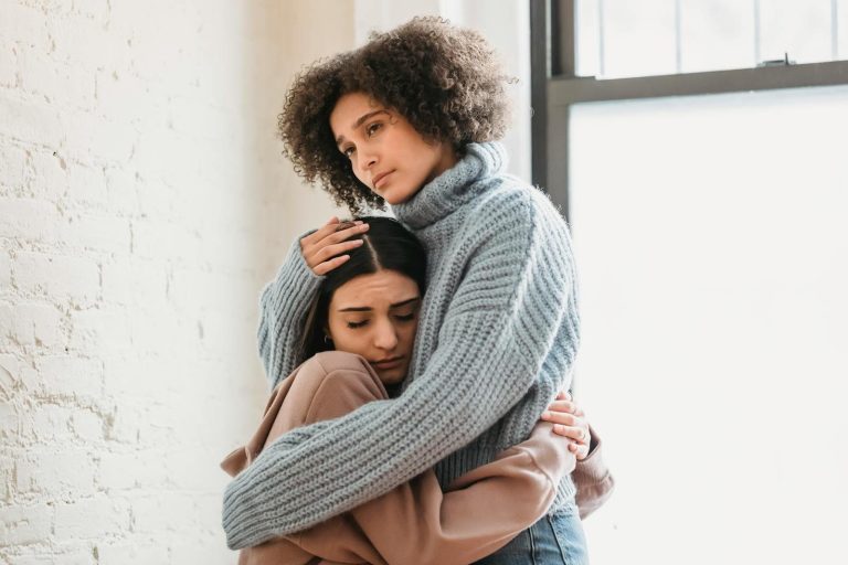 Frustrated African American woman embracing and comforting sad female friend while standing near window and wall in room at home